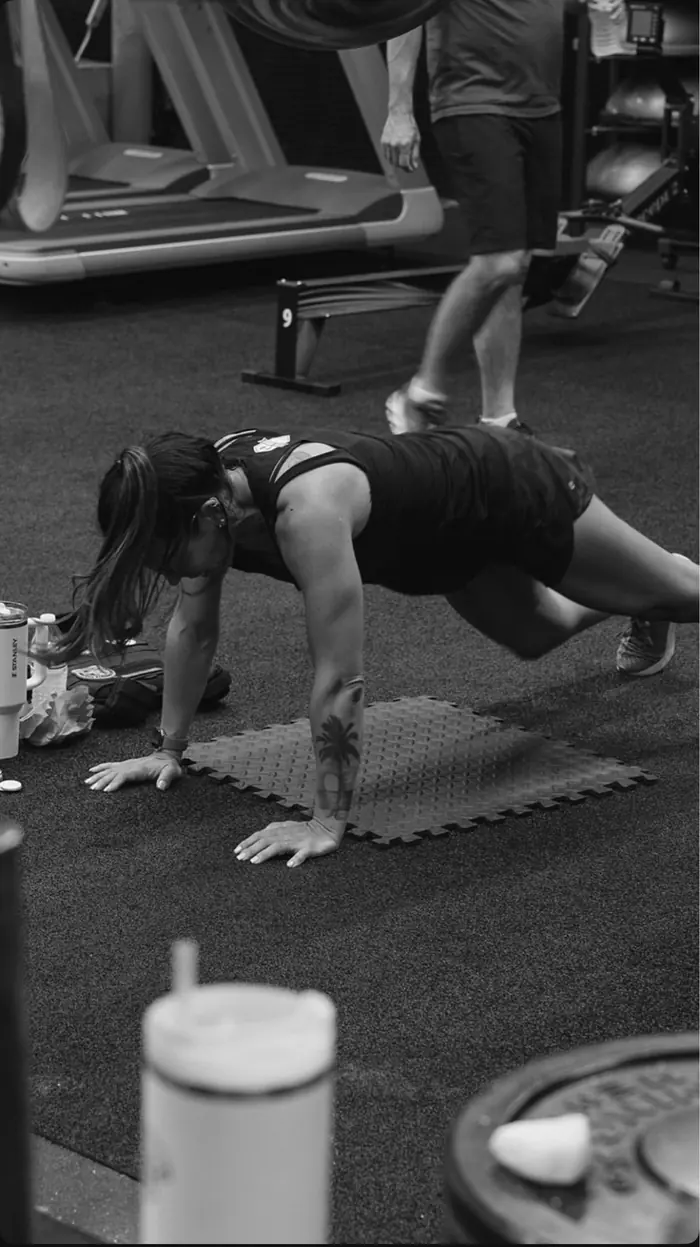 Black and white photo of a member holding a plank position on the gym floor while others train behind her.
