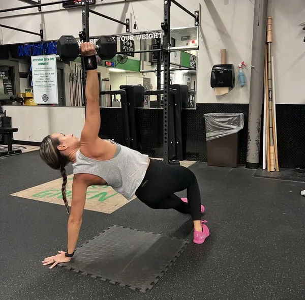 A member holding a side plank while pressing a dumbbell overhead in front of the rig.