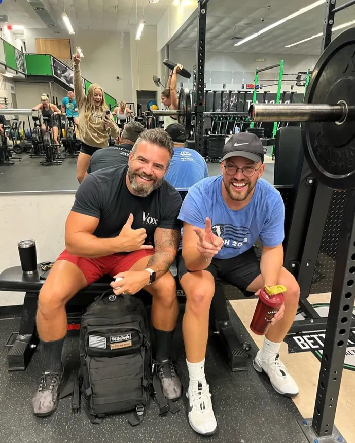 Two smiling members sitting on a bench beside a loaded barbell while a class trains behind them.